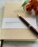 A beige linen hardcover journal with the title 'A Journal TOWARD TODAY' on the cover, accompanied by a pen and a ribbon bookmark.